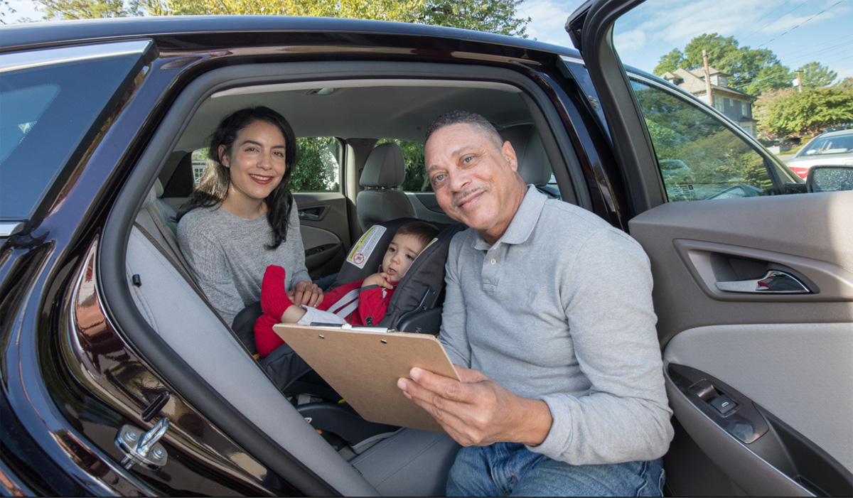 Mom with her baby sitting in the car.Male car seat technician has a clipboard in hand and is smiling at the camera.