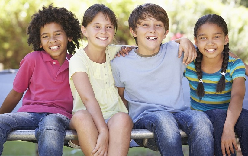 African American boy with pink polo, jeans, White grl with yellow top and shorts, White Boy with blue tee and jeans, Spanish-looking girl with blue striped top and jeans, with arms around each other smiling at the camera