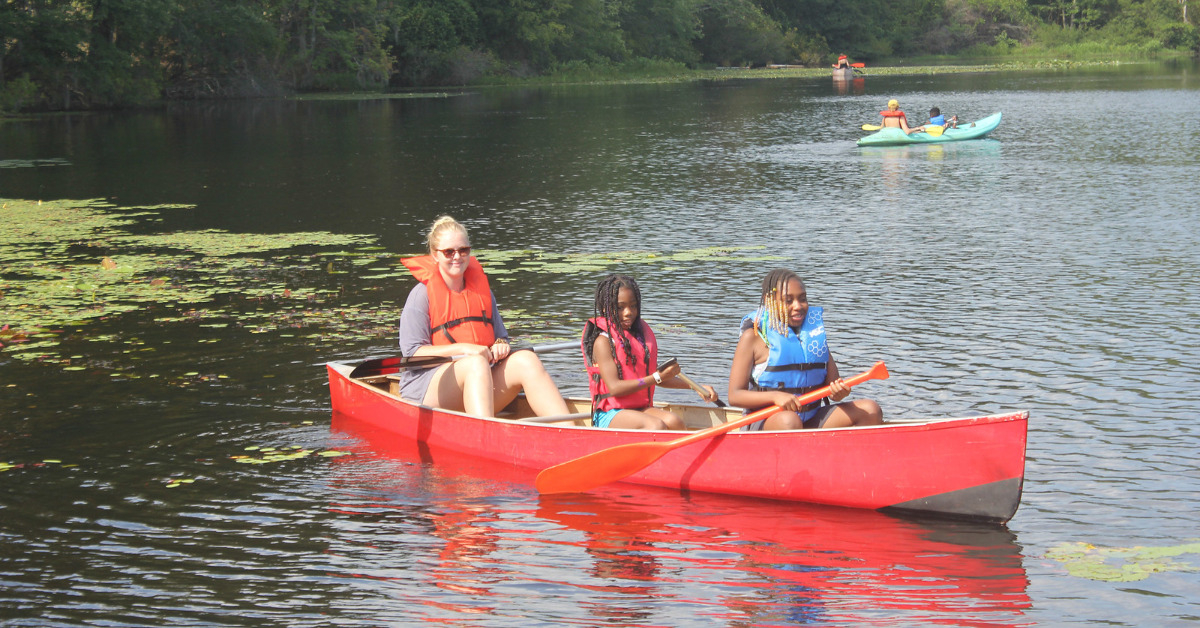 Two campers and one counselor in life jackets rowing in red canoe