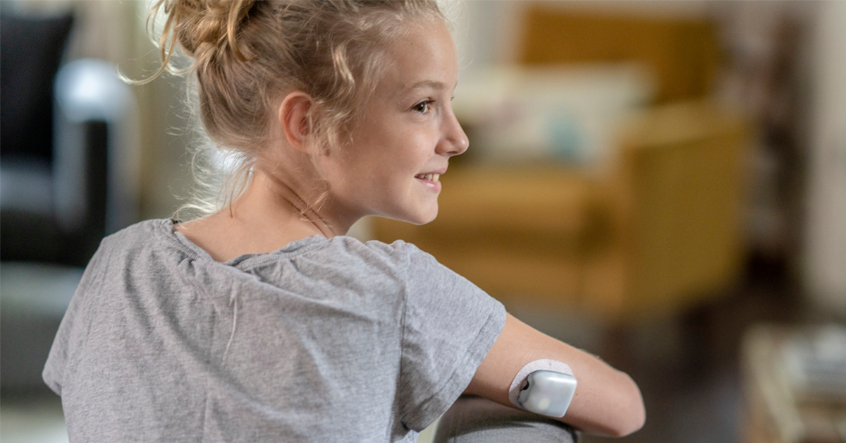 Young blonde girl in gray tee shirt turns around her right arm which is propped on the top of a chair showing her glucose monitor