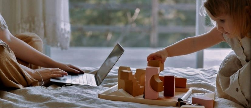 Young girl sitting on the bed with her mom whose typing on a laptop. The little girl is playing with pink wooden blocks