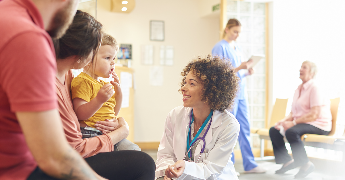 Family with young boy speaking with African American doctor with short curly hair. She is kneeling looking and smiling at the boy