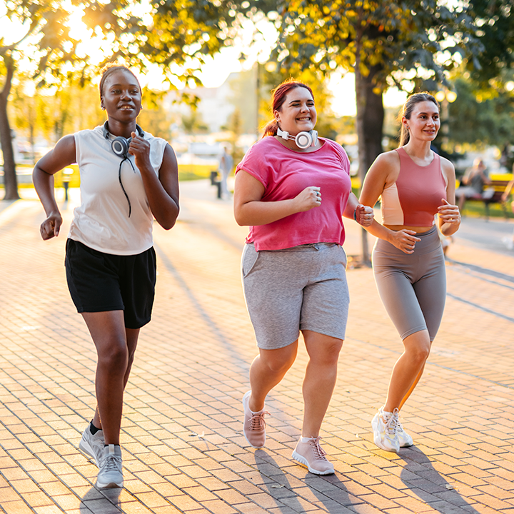 Three women jogging together on a brick sidewalk on a sunny morning. They all have varying shades of pink tank tops and gray biker shorts.