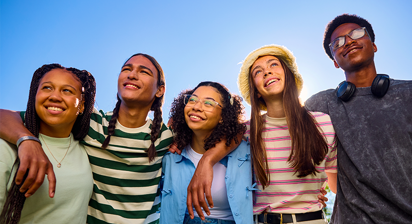 Diverse group of five teens with their arms around each other smiling and looking off into the distance.