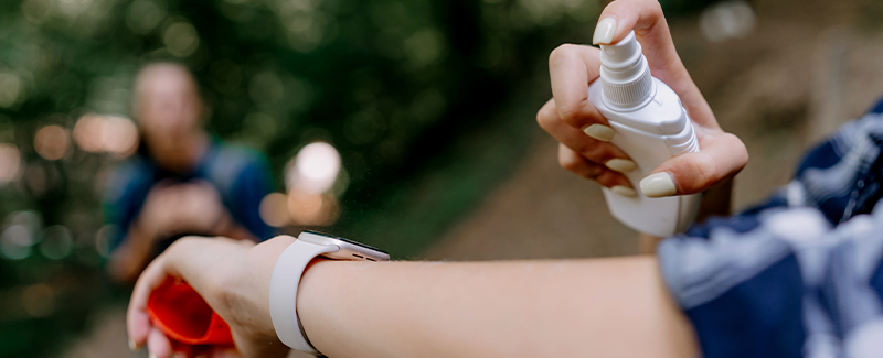 Woman with white nail polish and an Apple watch spraying mosquito repellant onto her forearm.